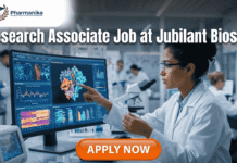 Research Associate Job at Jubilant Biosys –MPharm Candidates Apply Before the Deadline! Research Associate Job at Jubilant Biosys | M Pharm Candidates. A professional 1280x720 featured image for a "Research Associate Job at Jubilant Biosys." The scene features a female scientist in a white lab coat working in a bright, modern laboratory, pointing at a large monitor displaying colorful 3D molecular structures and research data. The Biotecnika logo is at the top center, and a prominent orange "Apply Now" button is at the bottom.